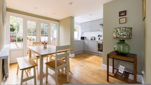 A kitchen with white cabinets, a dining table with chairs, a green vase on a side table, and a window with blinds. - The Thames Embrace 3 BR Garden House in Battersea (London)