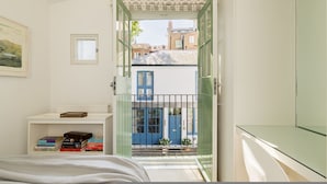 Ein Schlafzimmer mit einem Bett, einem Nachttisch mit Büchern, einem Fenster mit Blick auf Gebäude und einer Tür, die auf einen Balkon führt.