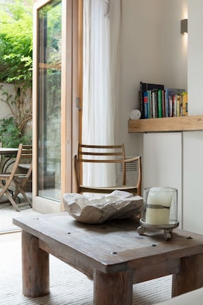 Ein Holztisch mit einer Steinschüssel und Kerze, ein Holzsessel, ein Bücherregal mit Büchern und eine Glastür, die in einen Garten führt.