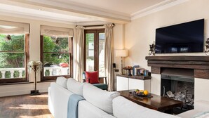 Ein Wohnzimmer mit einer Couch, einem Kamin, einem Fernseher und großen Fenstern mit Vorhängen.