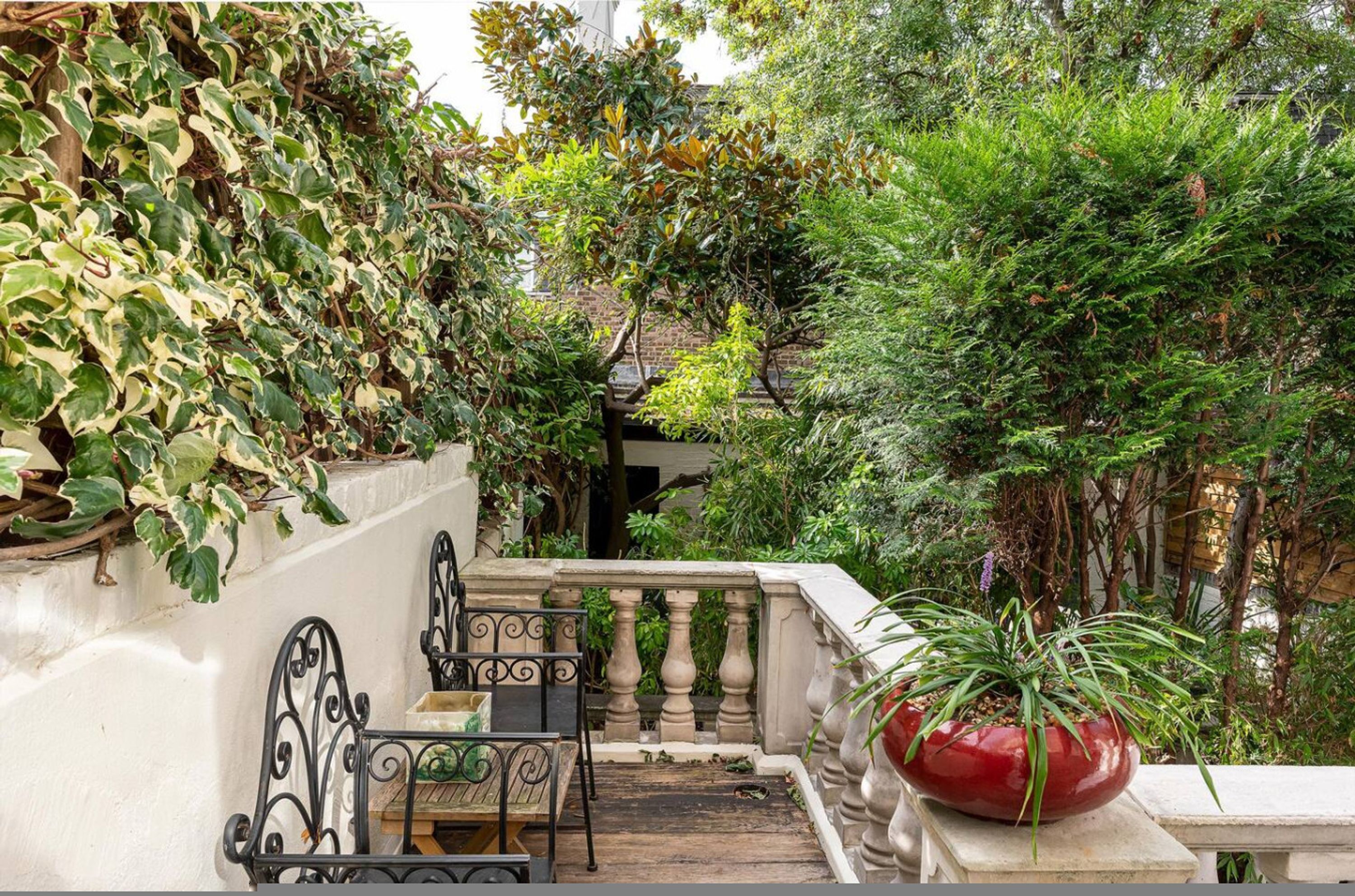 A balcony with a wrought iron chair, a red pot, and dense foliage.
