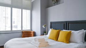 A neatly made bed with white linens and yellow pillows, a grey headboard, and a window with blinds. - Clapham Calm 2 Bedroom Flat With Garden (London)