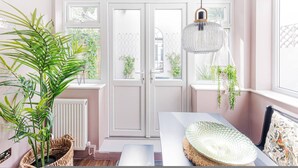 A room with a dining table, chairs, a hanging light fixture, and a large window with a view of greenery outside. - Clapham Calm 2 Bedroom Flat With Garden (London)