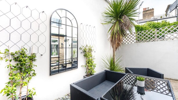 A patio with wicker furniture, potted plants, and a geometric wall design. - Clapham Calm 2 Bedroom Flat With Garden (London)