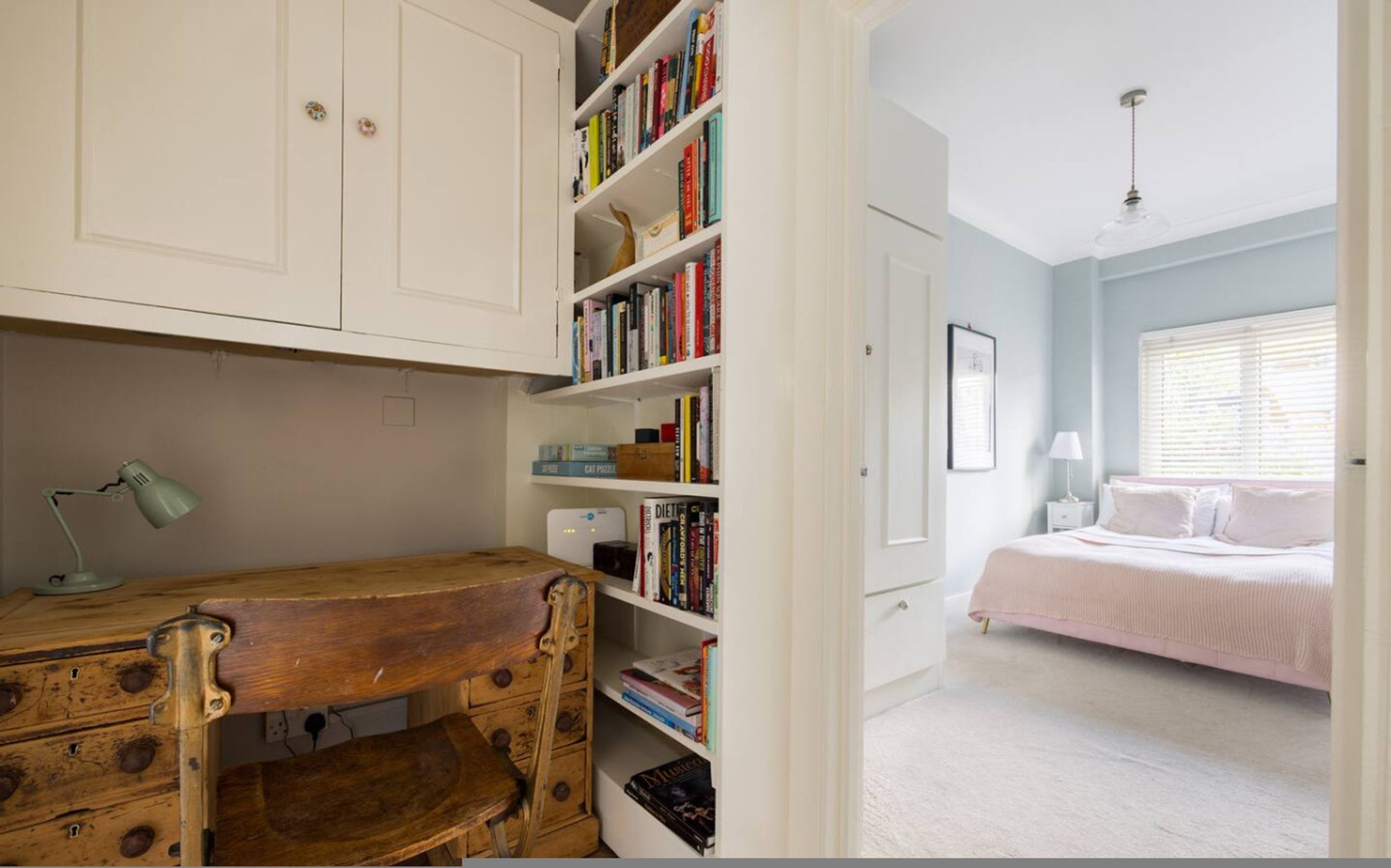A bedroom with a bed, a bedside table, a lamp, a window with blinds, and a bookshelf.