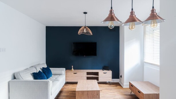 A modern living room with a dark blue accent wall, a flat-screen TV, a light-colored sofa with blue cushions, wooden coffee tables, and pendant lighting. - Stylish 1 BD in Notting Hill (London)