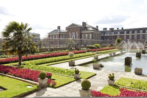 A formal garden with a fountain, neatly trimmed hedges, and a large building in the background. - Green History 3 Bedrooms in Kensington (London)