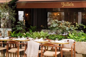 Ein Außenbereich mit Tischen und Stühlen, einem 'Julie's'-Sonnensegel und üppigem Grün.