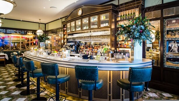 Un bar avec un sol à carreaux, des tabourets de bar, une grande horloge et un comptoir où sont disposées des bouteilles et des verres.