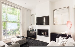 Ein modernes Wohnzimmer mit großem Fenster, Kamin, schwarzem Sofa, weißem Hocker und einer pinkfarbenen Stehlampe.