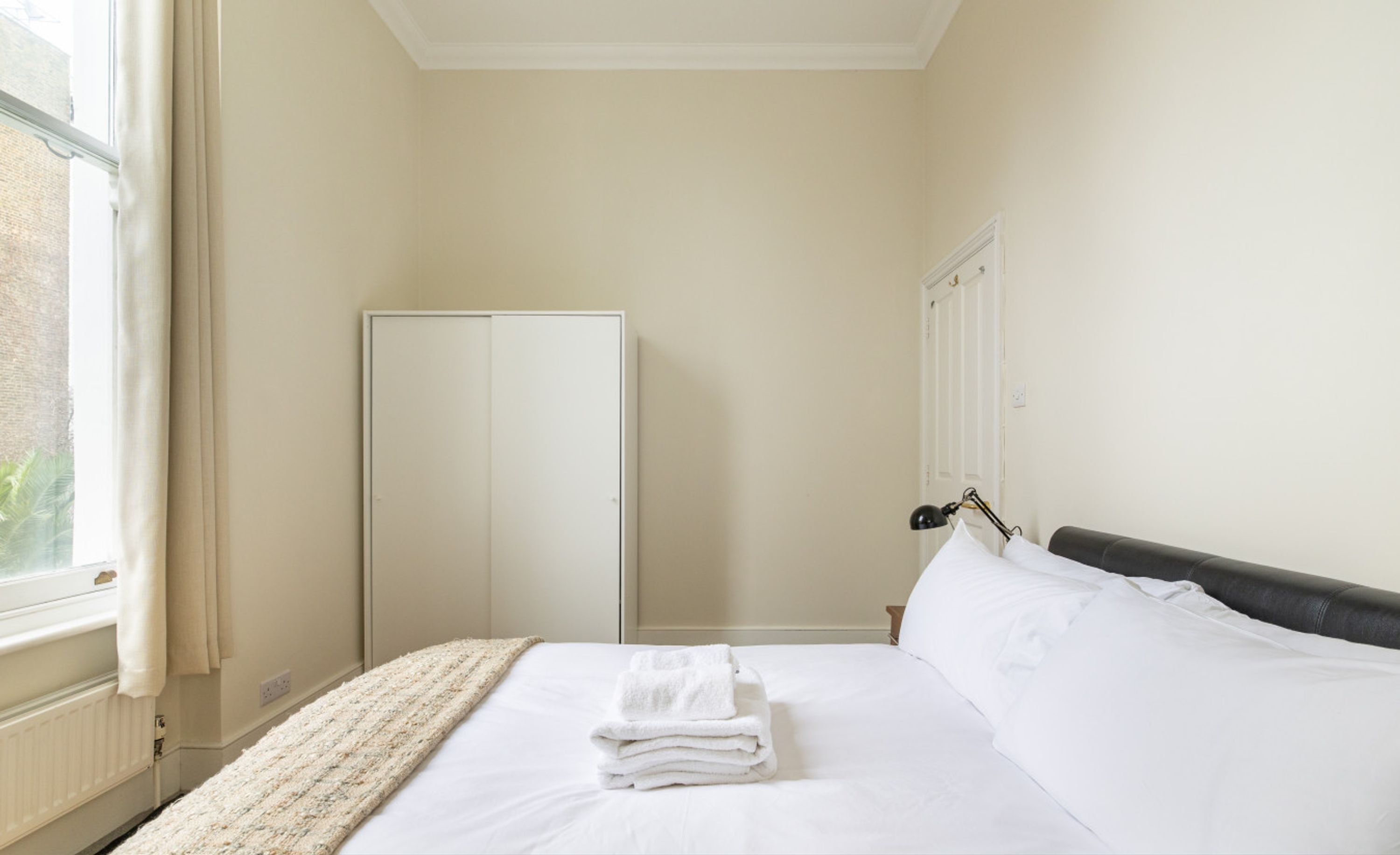 A hotel room with a bed, a window, a wardrobe, and a nightstand with a lamp.