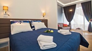 Ein Hotelzimmer mit einem großen Bett, zwei Nachttischlampen, einem Sessel, einem kleinen Tisch und Blick ins Freie.