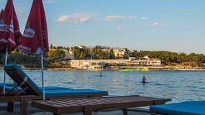 Ein Hotel am Wasser mit Liegestühlen und Sonnenschirmen, ein Strand im Hintergrund und Gebäude gegenüber dem Wasser.