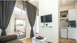Ein modernes Wohnzimmer mit einer Couch, einem kleinen Tisch, einem an der Wand montierten Fernseher und einer Küchenzeile mit Mikrowelle und Kühlschrank.