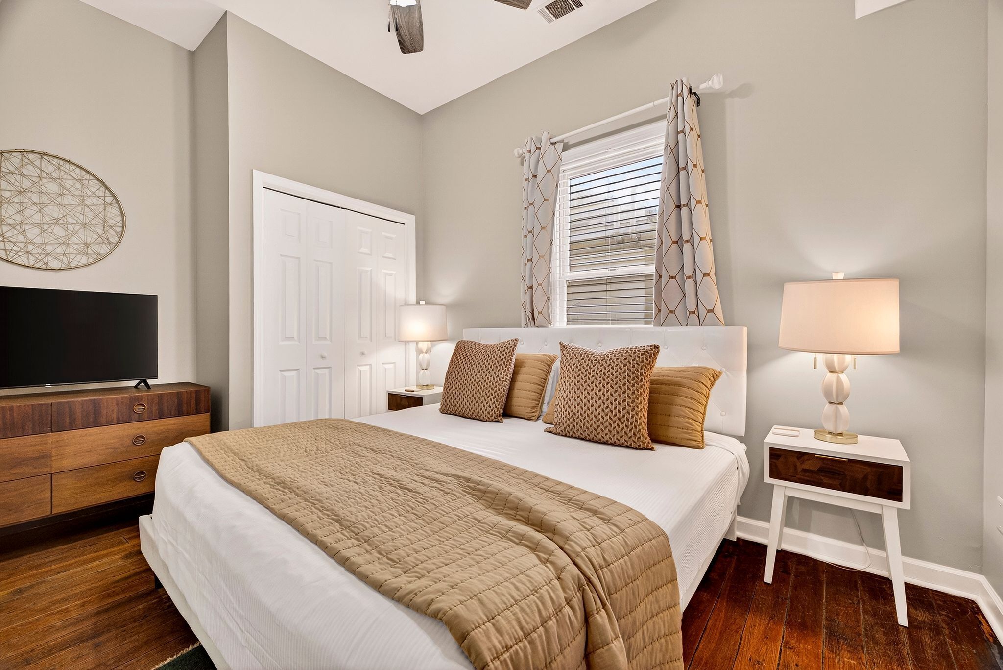 A bedroom with a bed, a dresser, a TV, a window with curtains, and a bedside lamp.