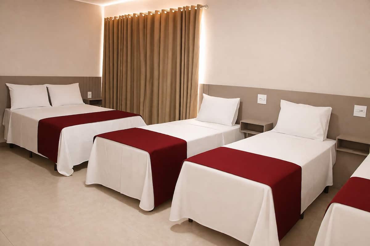 A hotel room with four single beds, each with a white bedspread and a red blanket. There is a large window with beige curtains, and the room has a neutral color scheme.