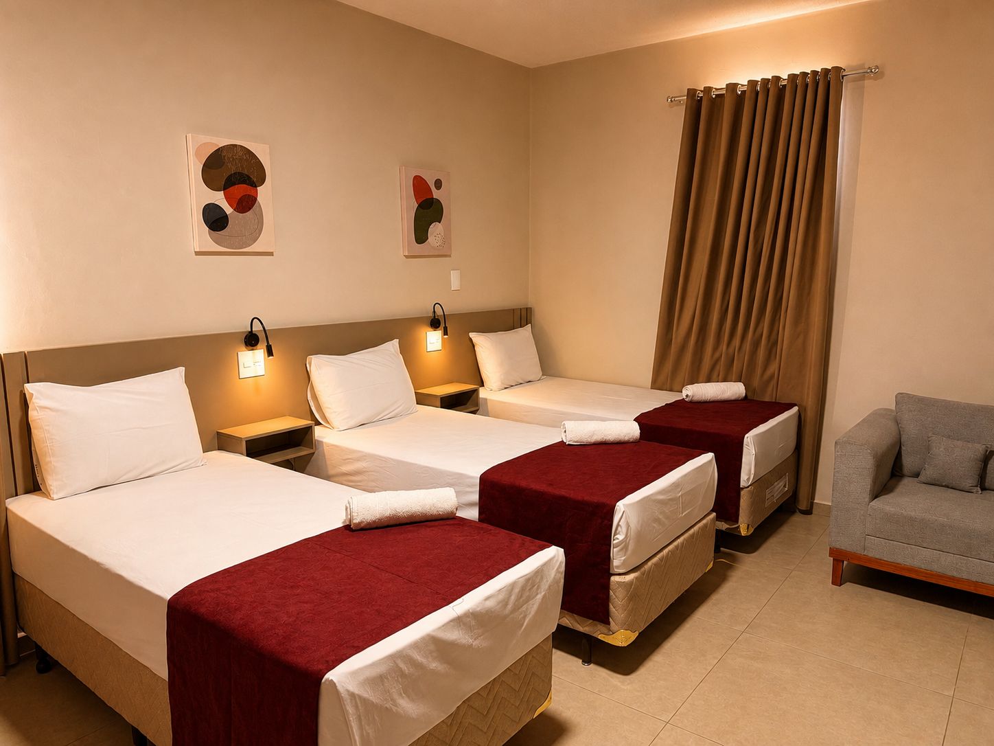 Quarto de hotel com três camas, um sofá cinzento e duas luminárias fixadas na parede.