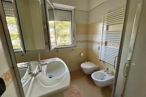 A bathroom with a sink, toilet, bidet, and a window with blinds. - Homiday - Raffaella, Three Bedroom Apartment with Balcony (Alba Adriatica)