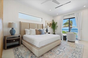 A modern bedroom with a large bed, bedside tables, a chair, and a view of the ocean.