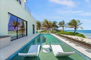 A pool with two lounge chairs, a clear view of the ocean, and palm trees.
