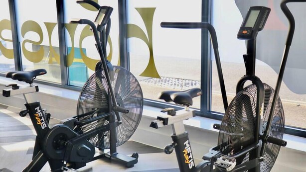 Tre motionscyklar med fläktar, en digital display och ett fönster med utsikt över stranden.