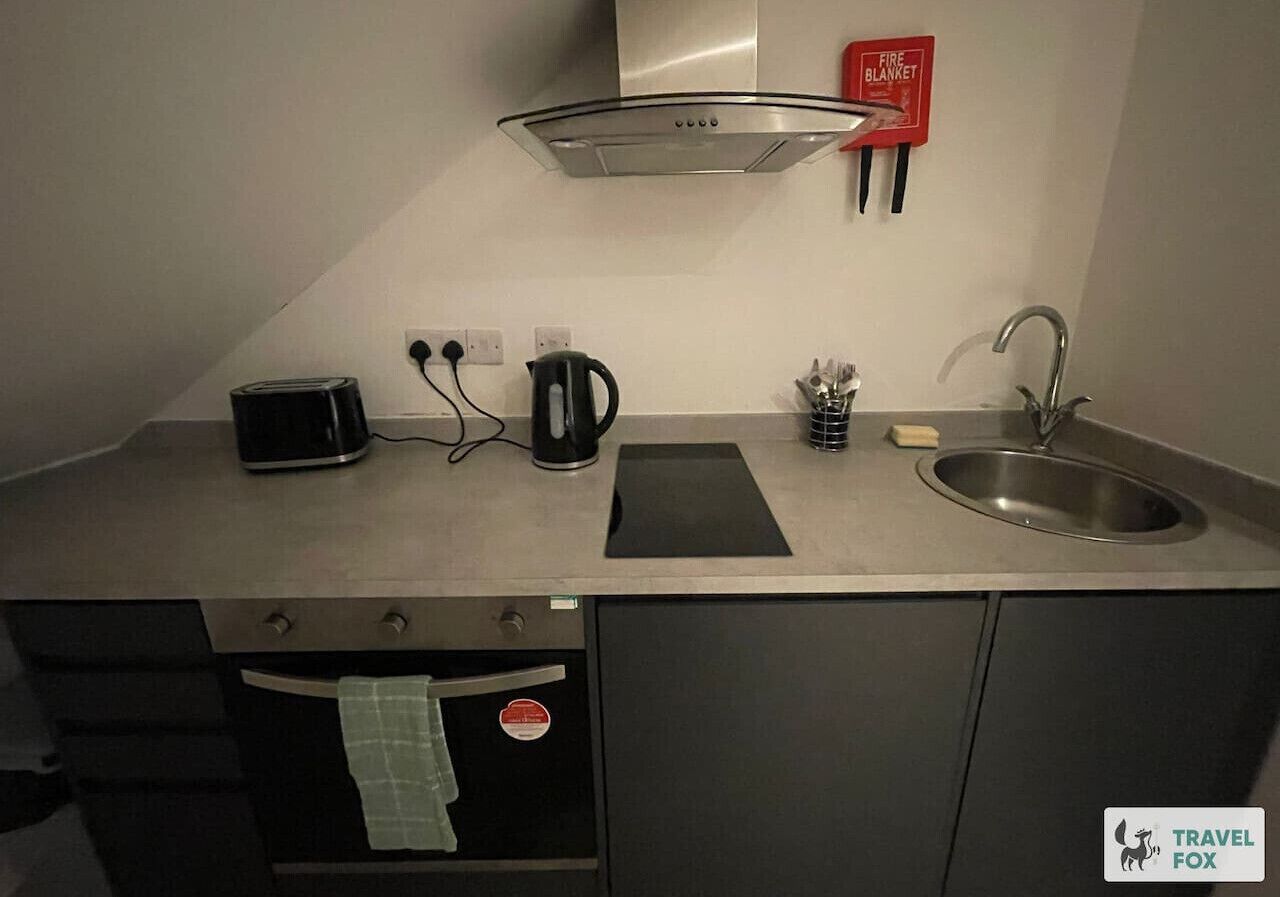 A compact kitchenette with a stove, kettle, toaster, sink, and a fire blanket.