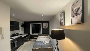 Ein kleines Hotelzimmer mit Bett, Nachttisch, Lampe, einer Küchenzeile und einer kleinen Sitzecke.