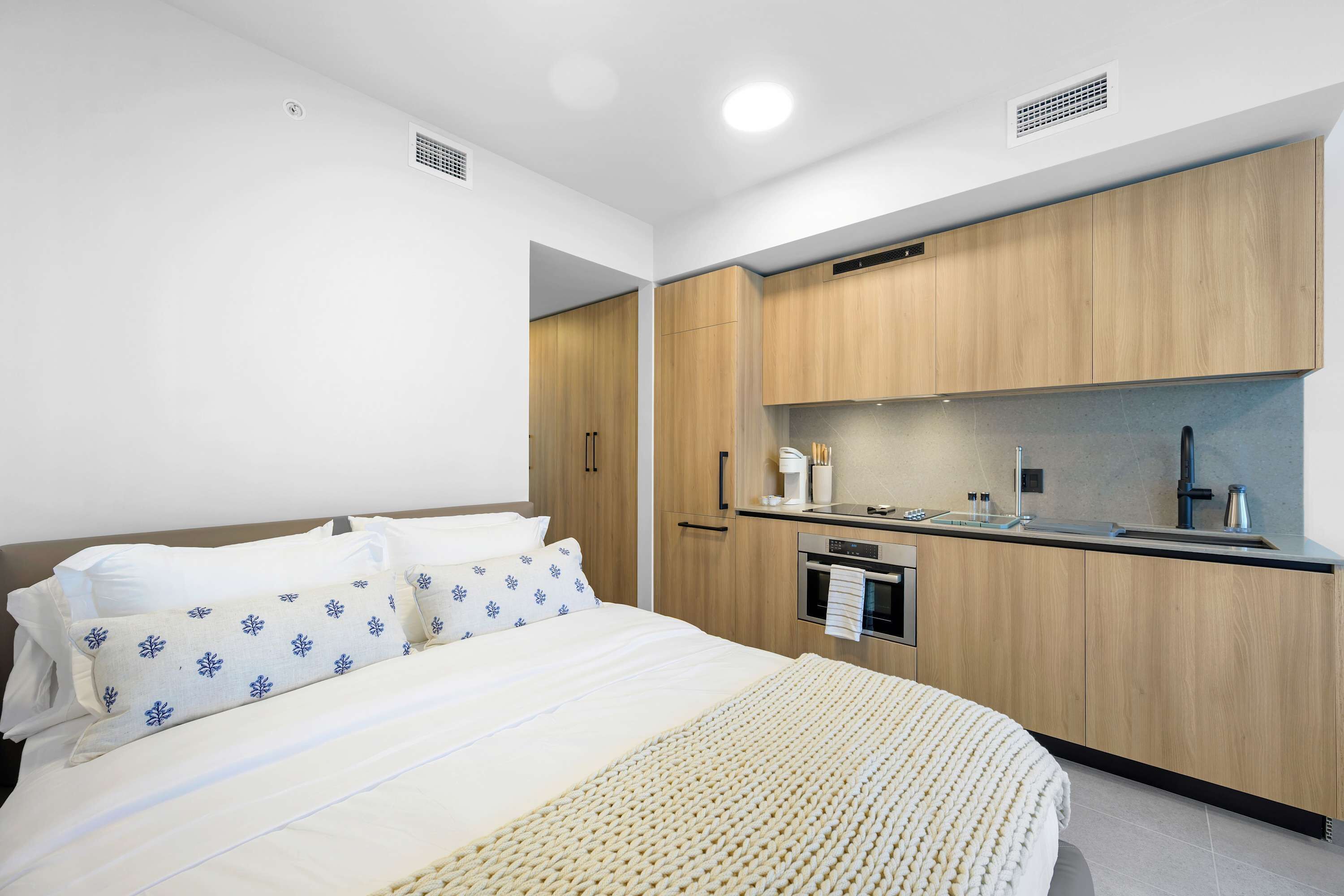 A modern hotel room with a bed, a kitchenette, and a bathroom.