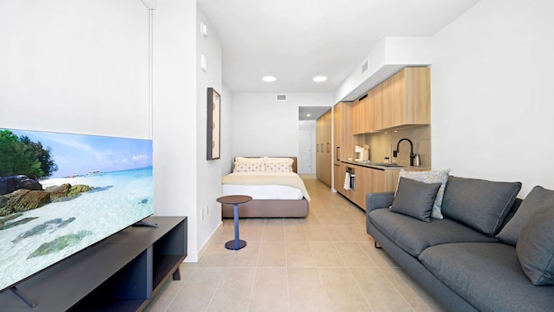 A modern hotel room with a TV displaying a beach scene, a sofa, a small table, and a kitchenette. - NOK Urban Studio in Prime Miami Location (Miami)