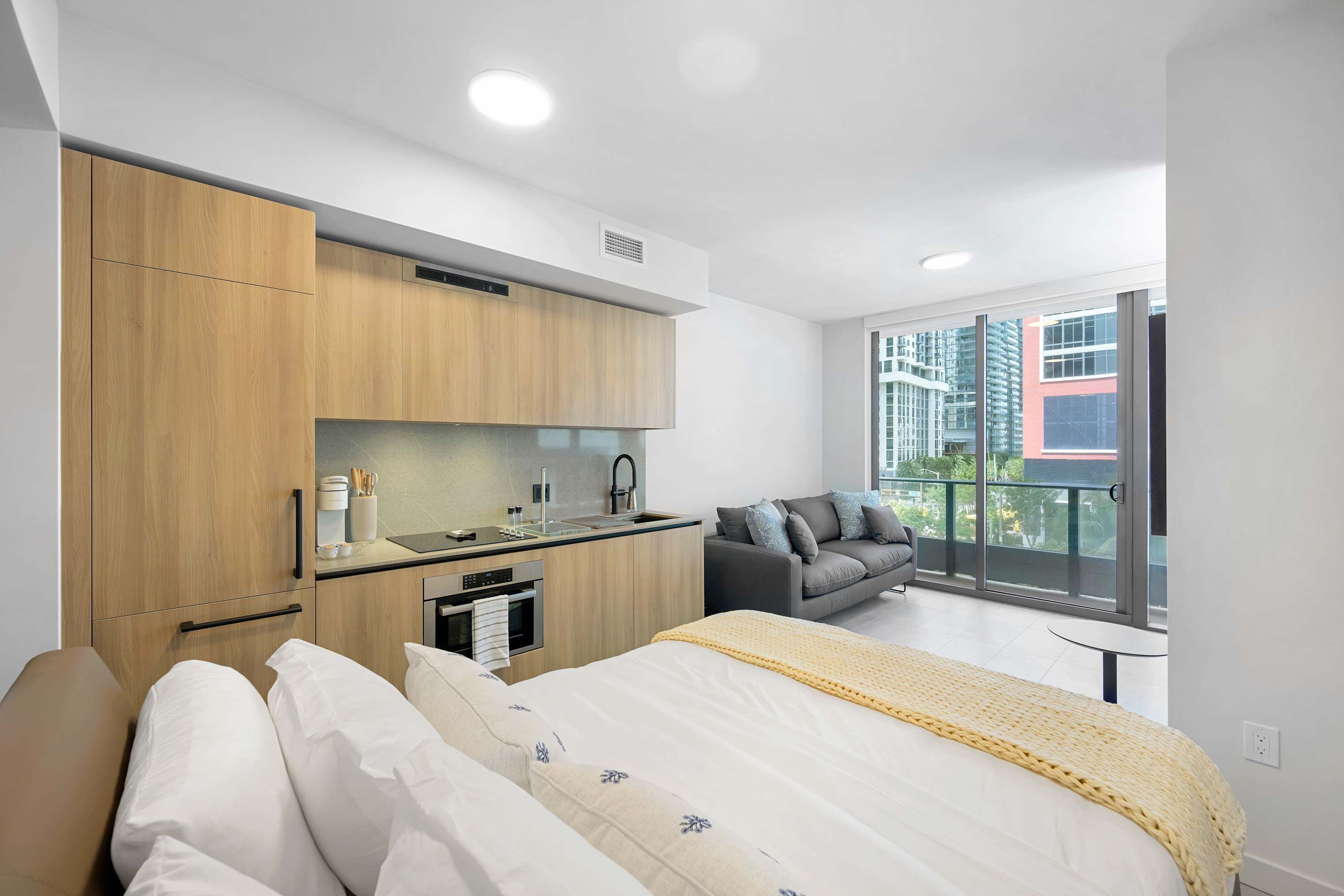 Una habitación de hotel moderna con kitchenette, una cama con manta amarilla, un sofá gris y un balcón con vista a la ciudad.
