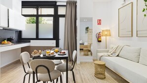 En moderne stue med sofa, spisebord med stole, et køkkenområde med et vindue og en lampe.