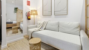 En moderne stue med sofa, lampe, lille rundt bord og et spejl der reflekterer rummet.