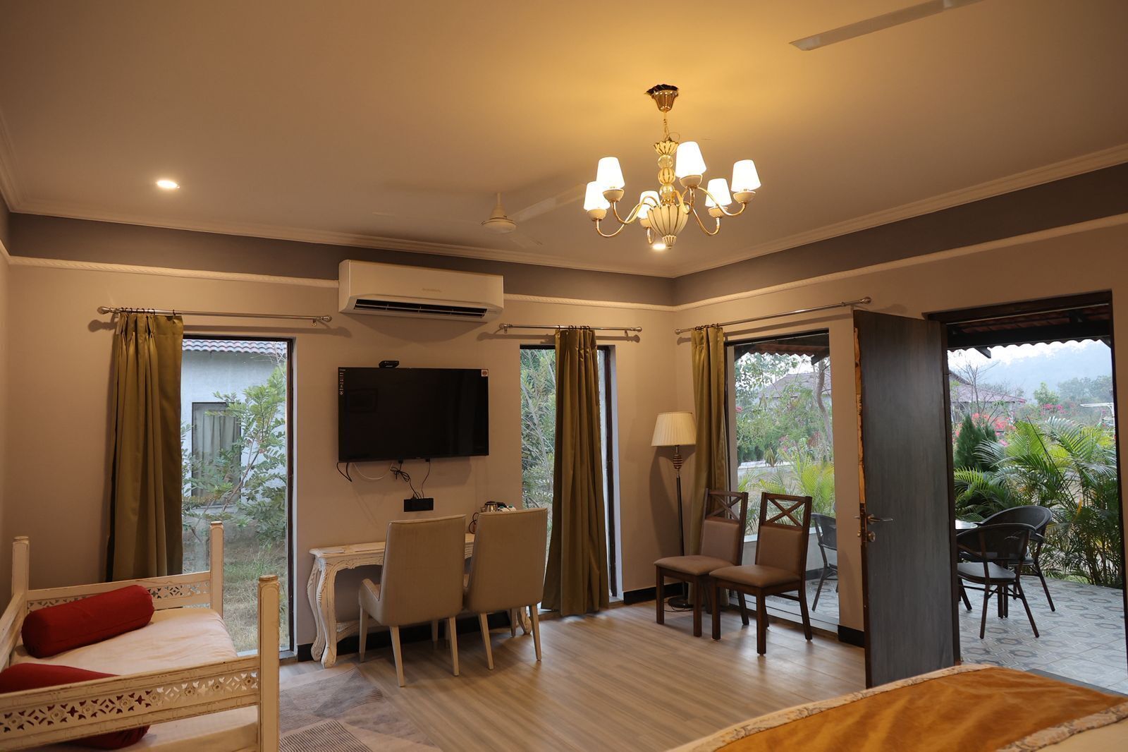 Habitación de hotel con cama, televisor, escritorio con sillas, candelabro y vista a la vegetación.