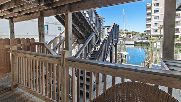 Une terrasse en bois dotée d’une rambarde, d’un escalier et offrant une vue sur une marina avec des bateaux et des bâtiments.