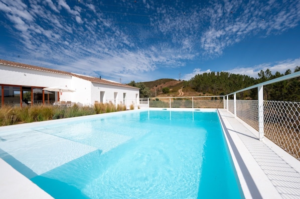 Une piscine avec un bâtiment blanc en arrière-plan, entourée d’une clôture et de végétation.