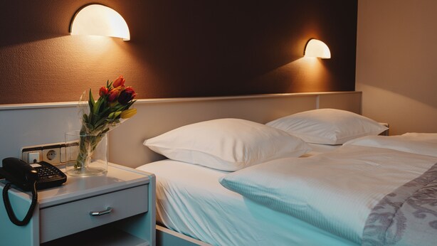 A hotel room with a bed, bedside table, lamp, and a vase with flowers. - Hotel Casa56 (Weiswampach)
