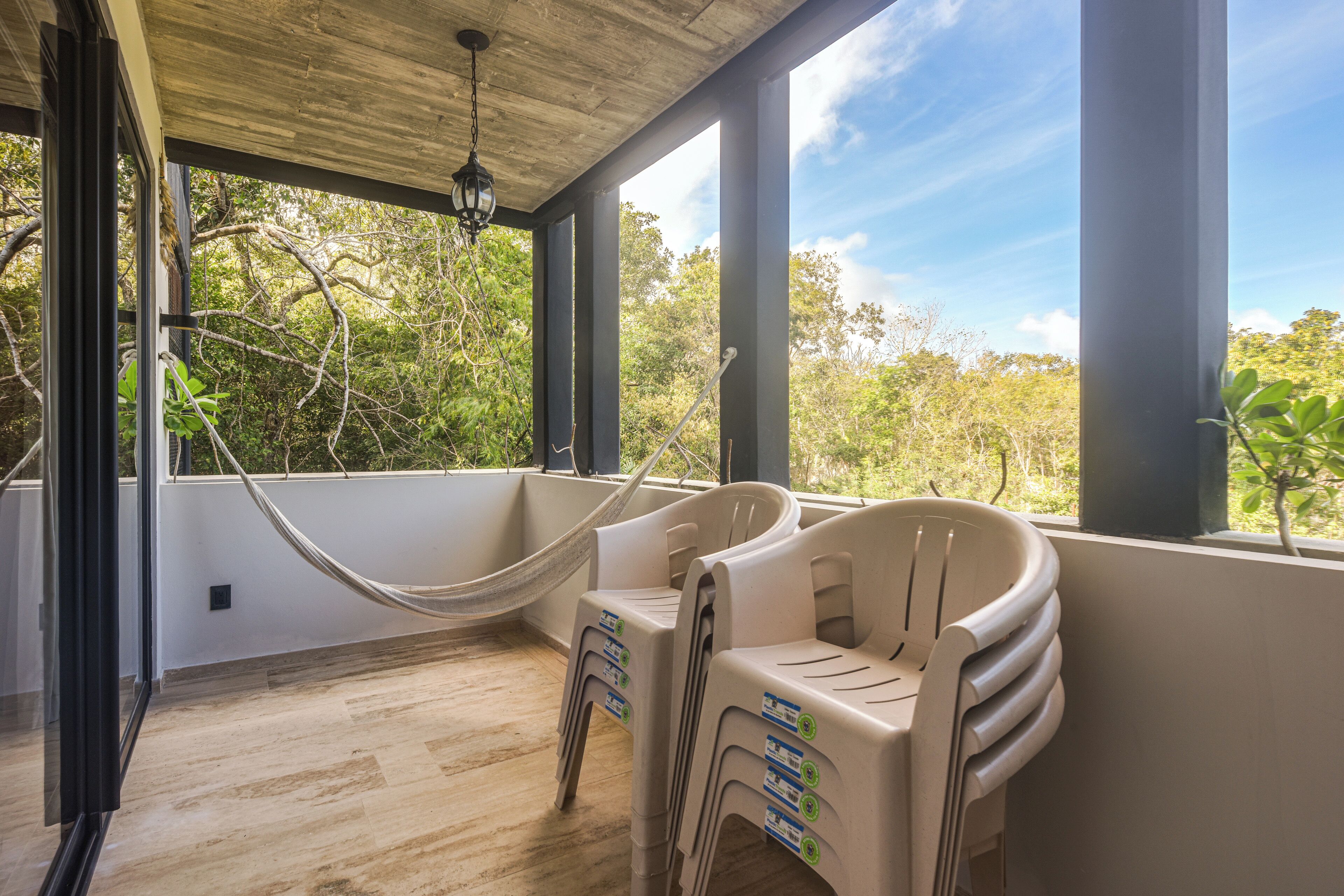 A room with a hammock, stacked chairs, and large windows overlooking trees.