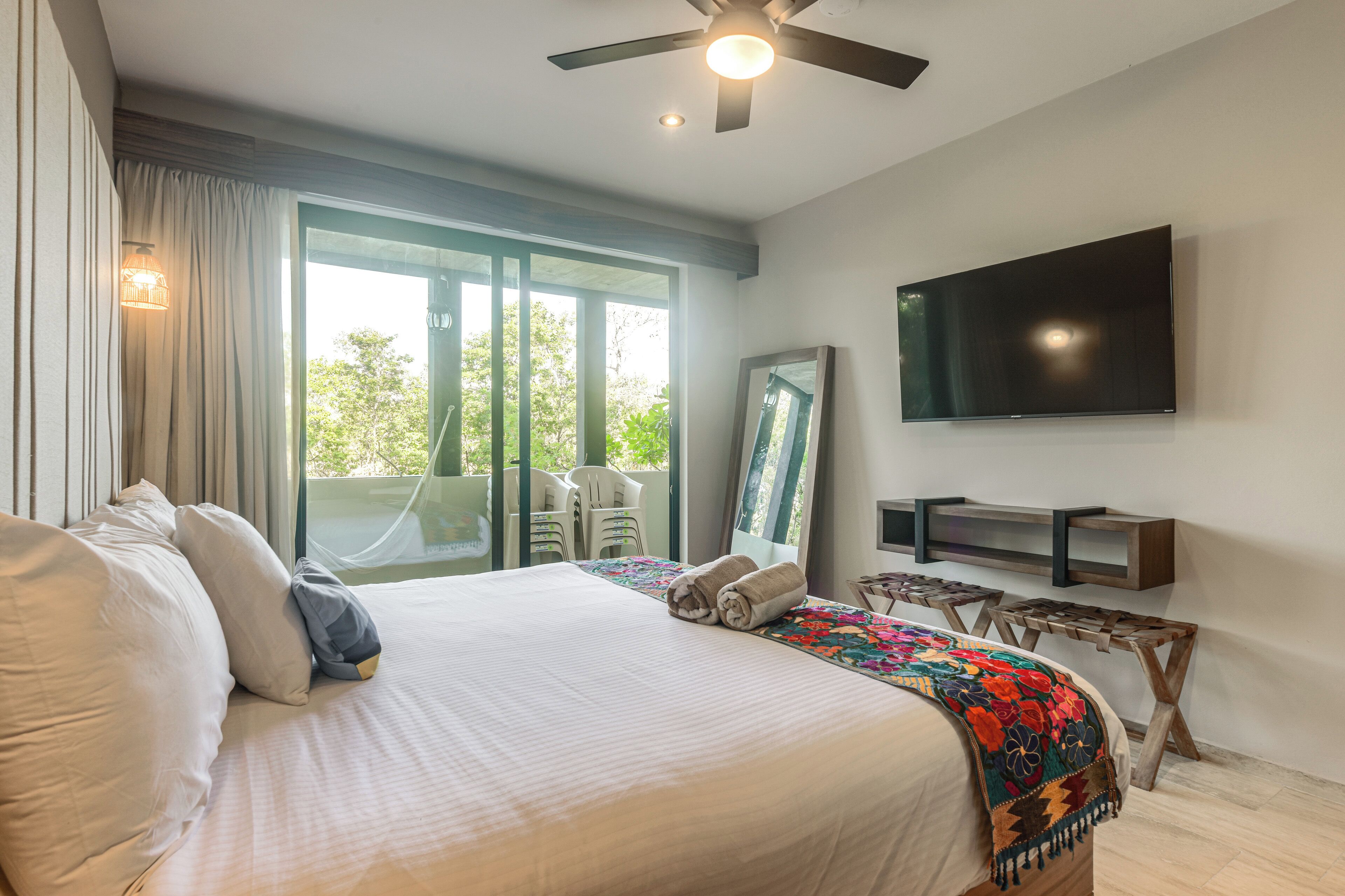 Une chambre d’hôtel dotée d’un grand lit, d’un ventilateur de plafond, d’un téléviseur à écran plat, d’un balcon avec des chaises et d’une grande fenêtre.