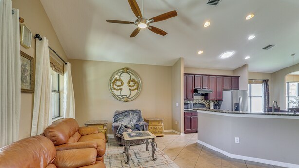 A living room with a leather sofa, a patterned rug, a decorative wall piece, a ceiling fan, and a kitchen area with cabinets and appliances. - Extended Stay in Tampa Area! Spacious Home w/ Yard (Riverview)