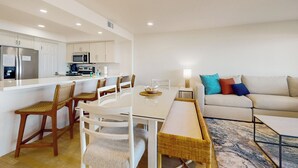 A modern kitchen with white cabinets, a refrigerator, and a microwave. There is a dining area with a white table and chairs, and a sofa with colorful pillows. - Sweeping View, Luxe Perks: Suntide III - Unit 708 (South Padre Island)