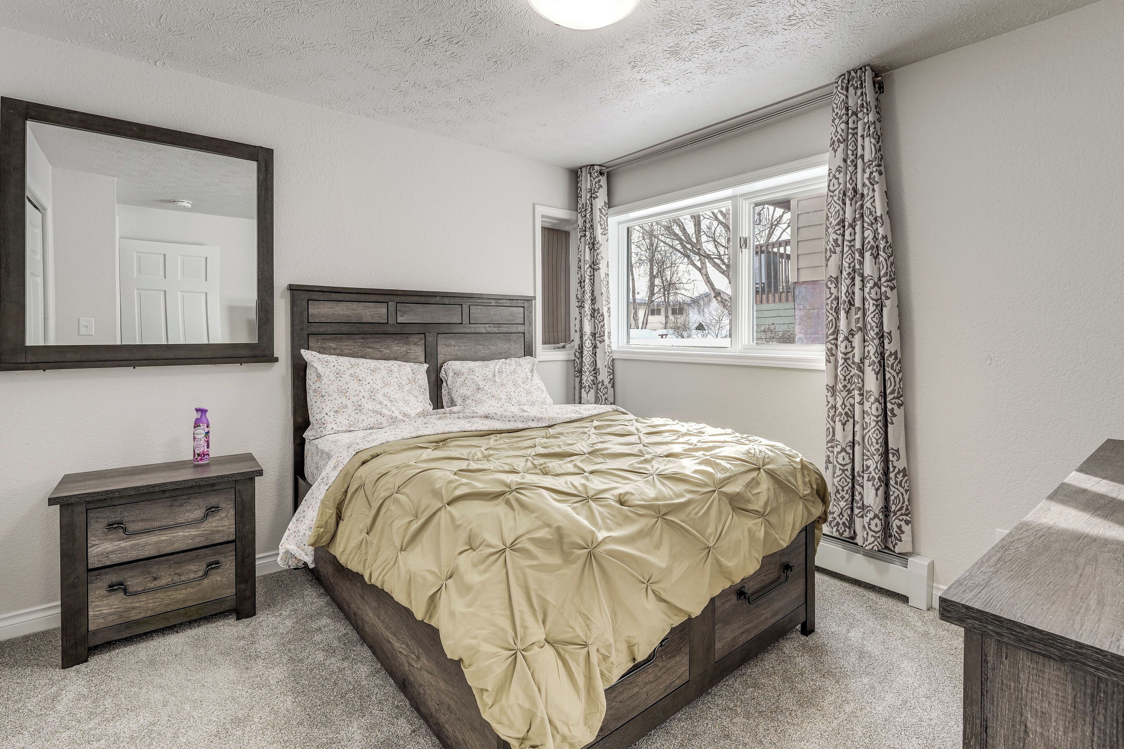A bedroom with a bed, a nightstand, a dresser, a window with curtains, and a mirror.