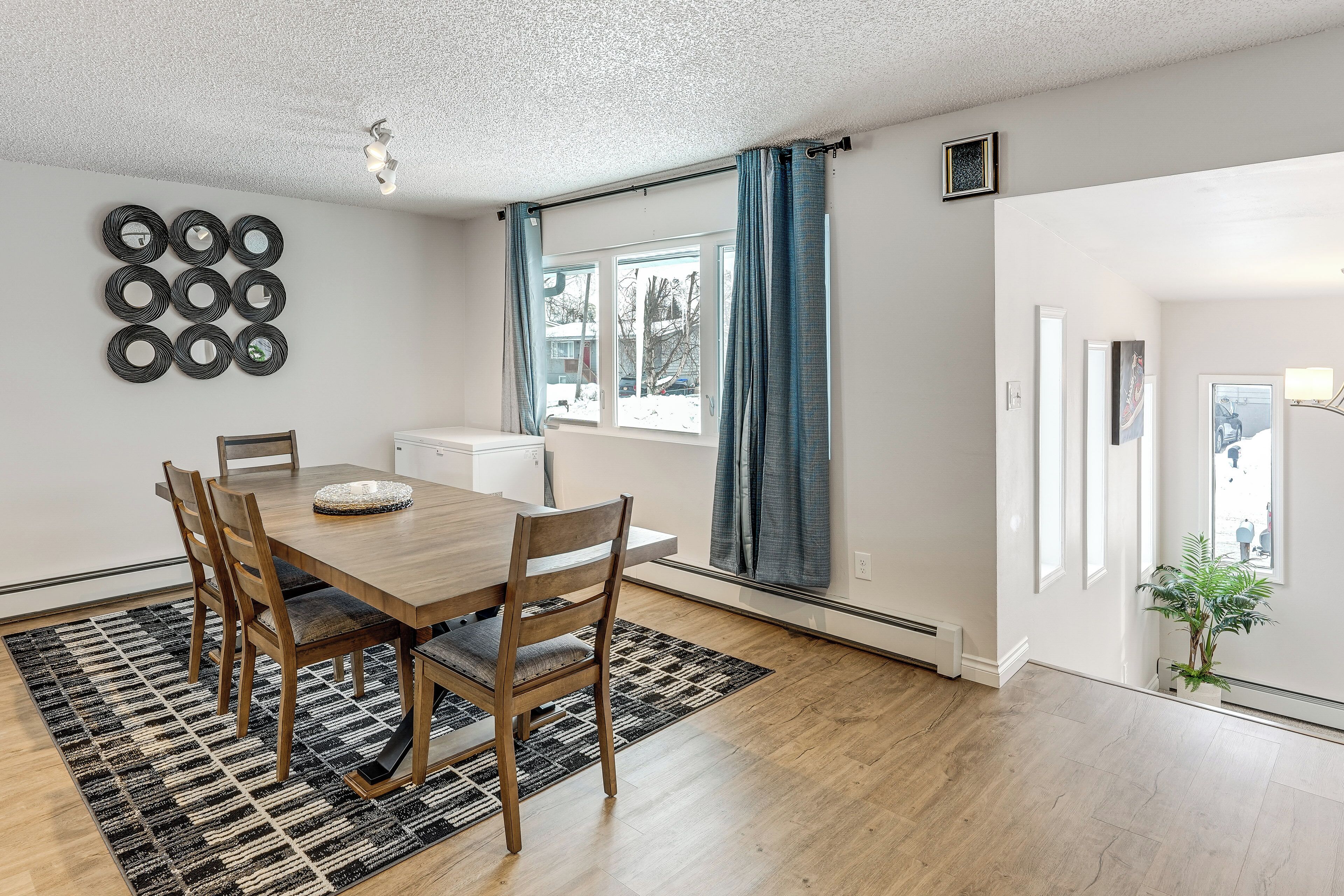 Un coin repas comprenant une table en bois, quatre chaises, un tapis à motifs, un miroir fixé au mur et une fenêtre avec des rideaux.