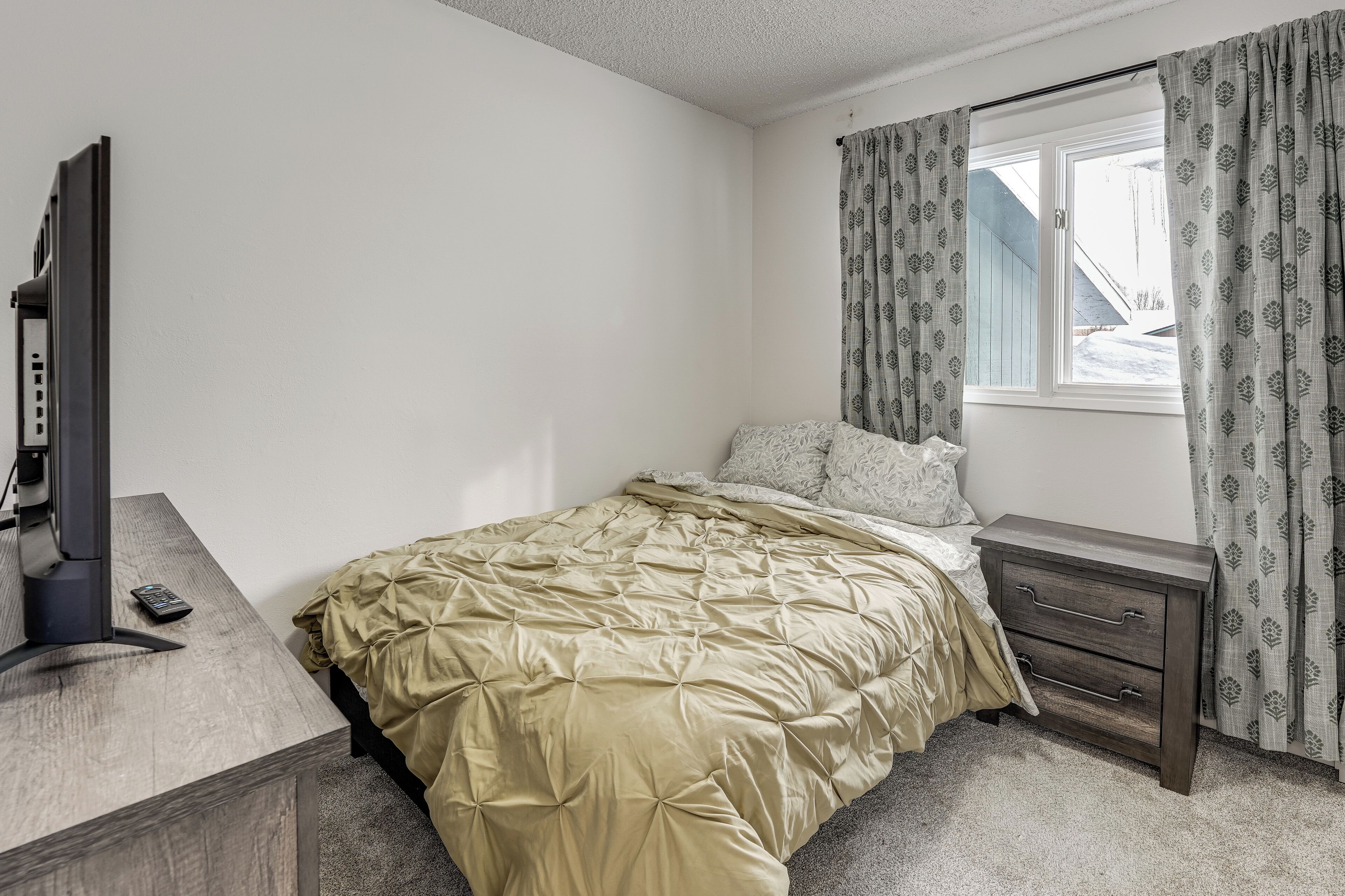 A bedroom with a bed, a bedside table, a TV, and a window with curtains.