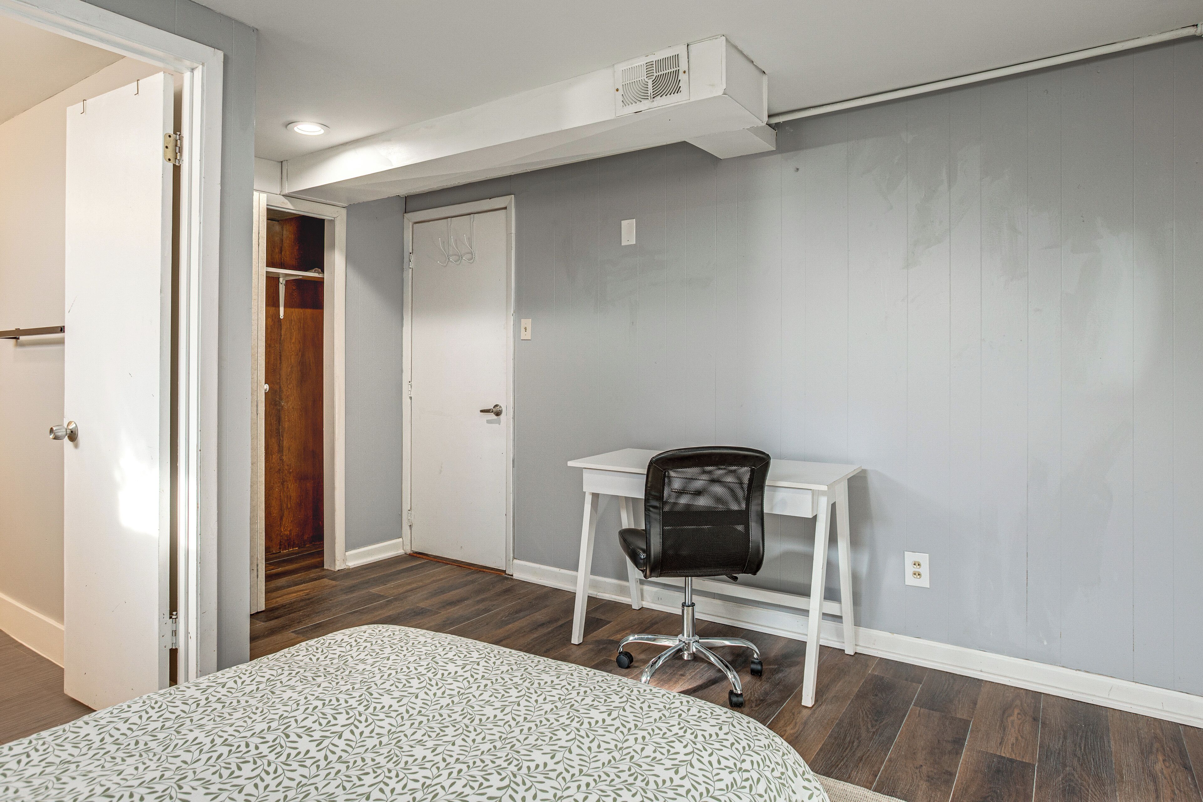 A bedroom with a bed, a desk, and a chair. There is a closet with a door, a white wall, and a ceiling vent.