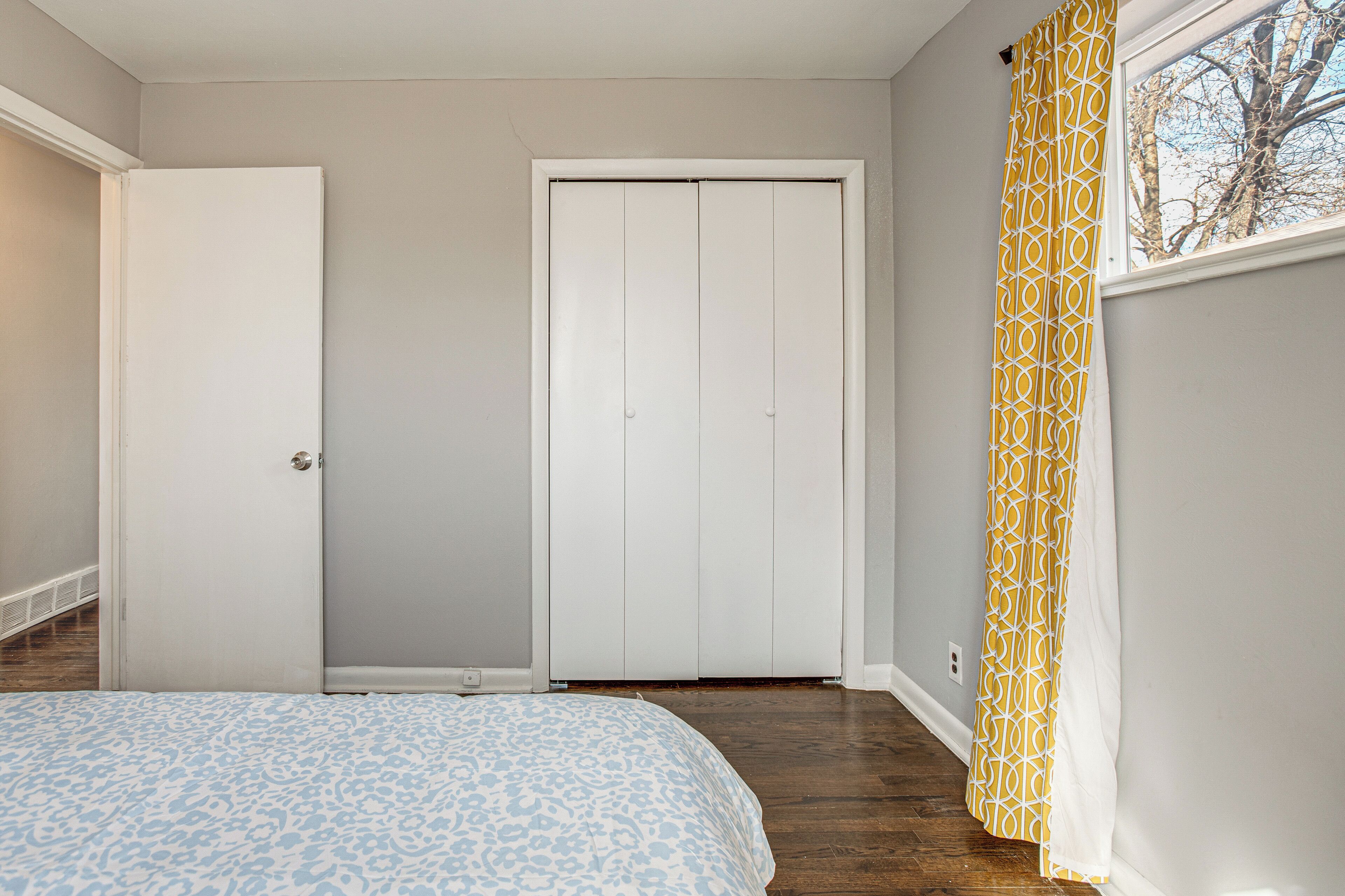 A bedroom with a bed, a closed door, a window with yellow curtains, and a closet.