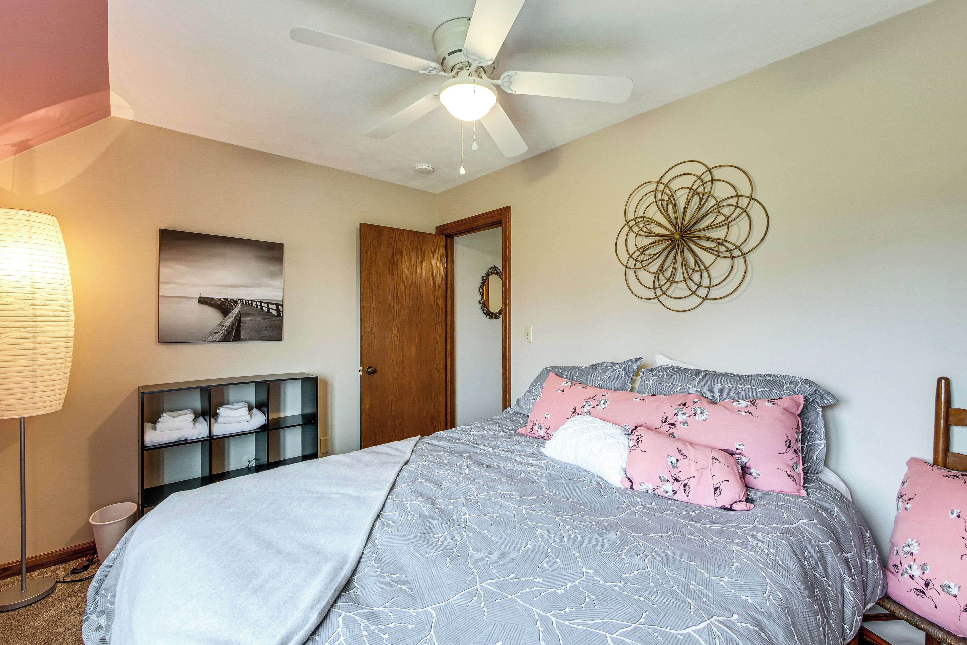 A bedroom with a bed, a ceiling fan, a wall-mounted artwork, a wooden door, and a bedside table with a lamp.