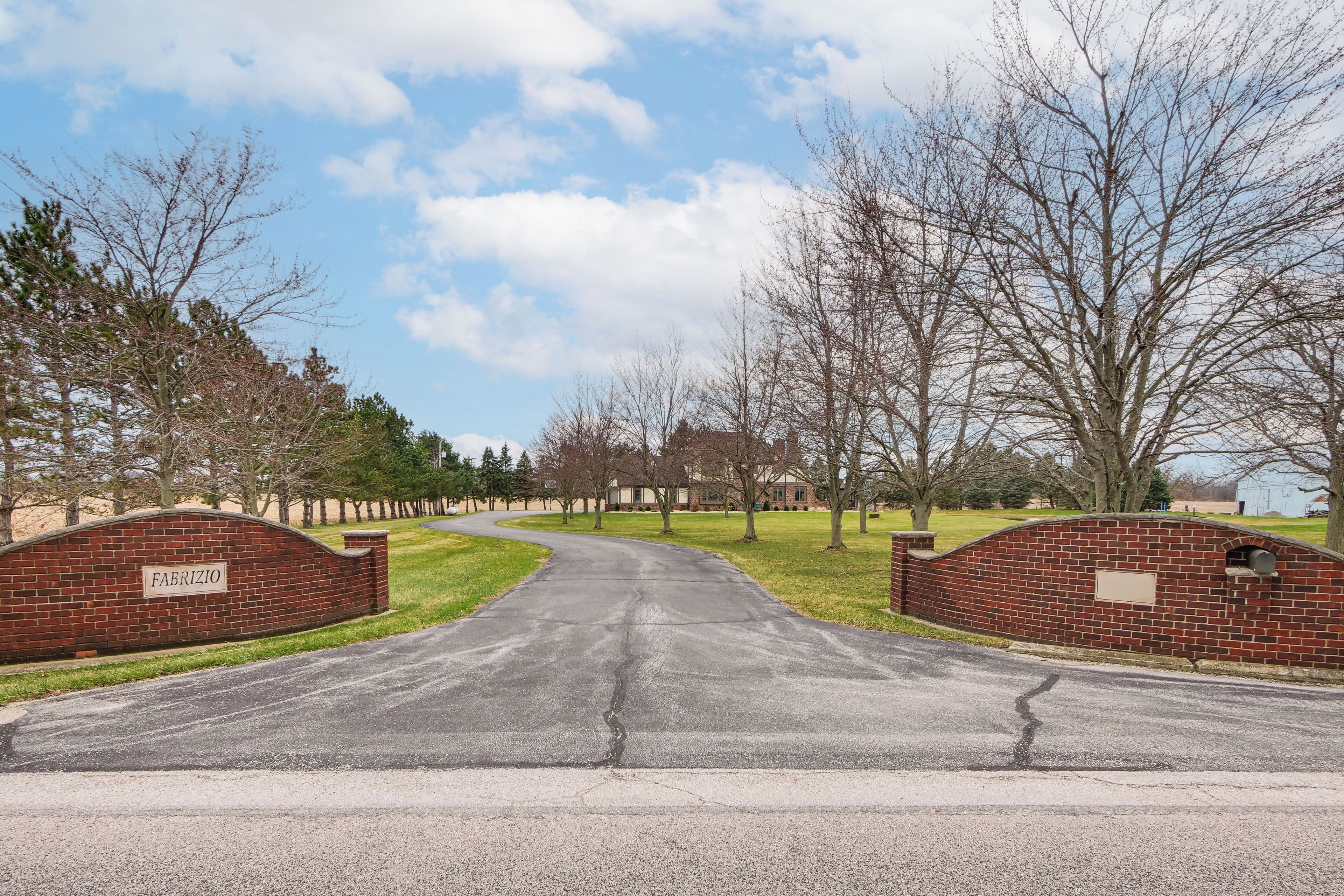 A paved driveway leading to a brick wall with a sign, surrounded by trees and a clear sky.