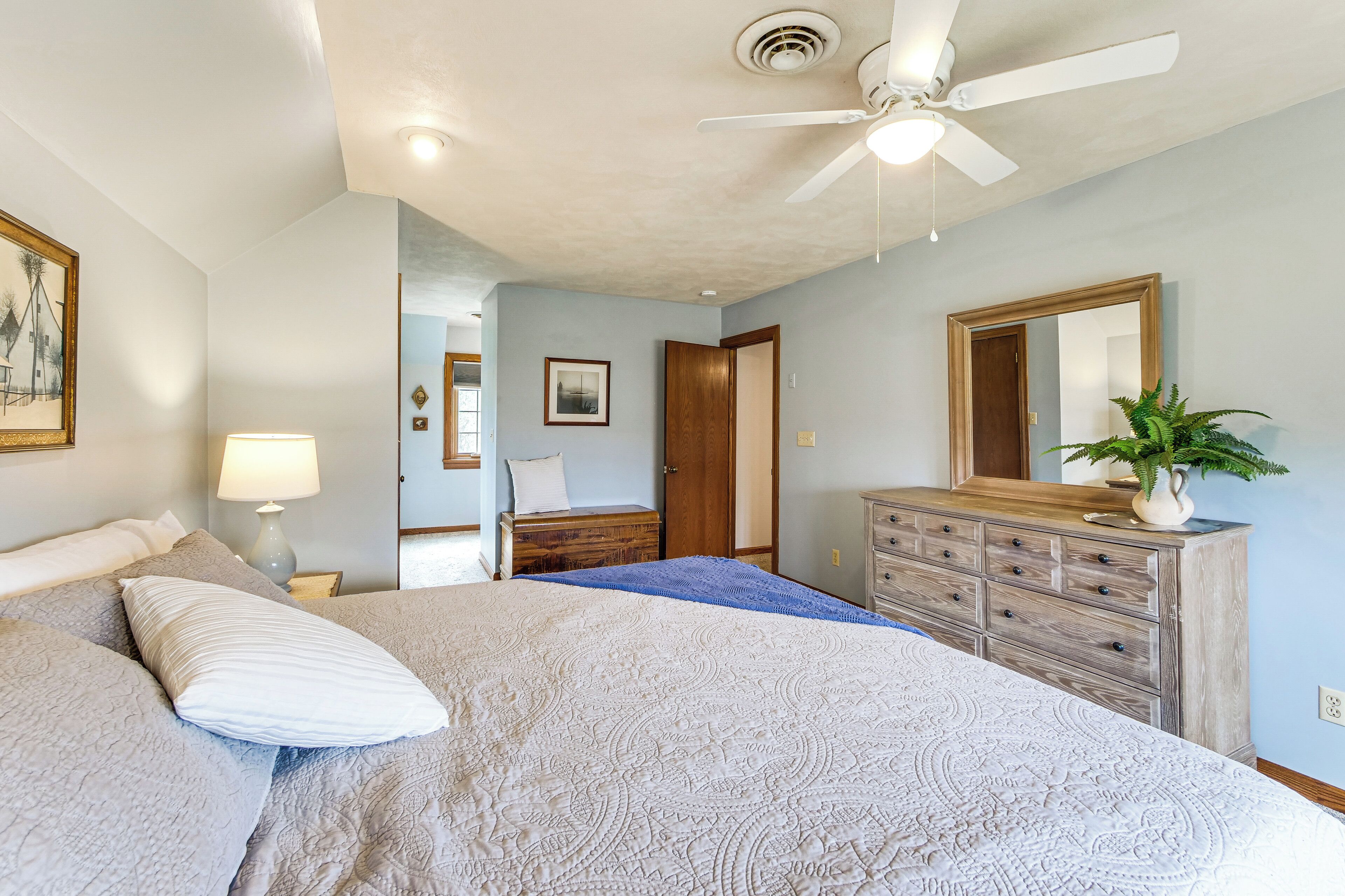 Une chambre à coucher comprenant un lit, une commode, un ventilateur de plafond et une porte donnant sur une autre pièce.