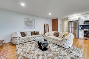 Ein modernes Wohnzimmer mit einem Glastisch, zwei weißen Sofas mit gemusterten Kissen, einer Wand in Holzoptik und einer Küchenzeile mit Edelstahlgeräten.