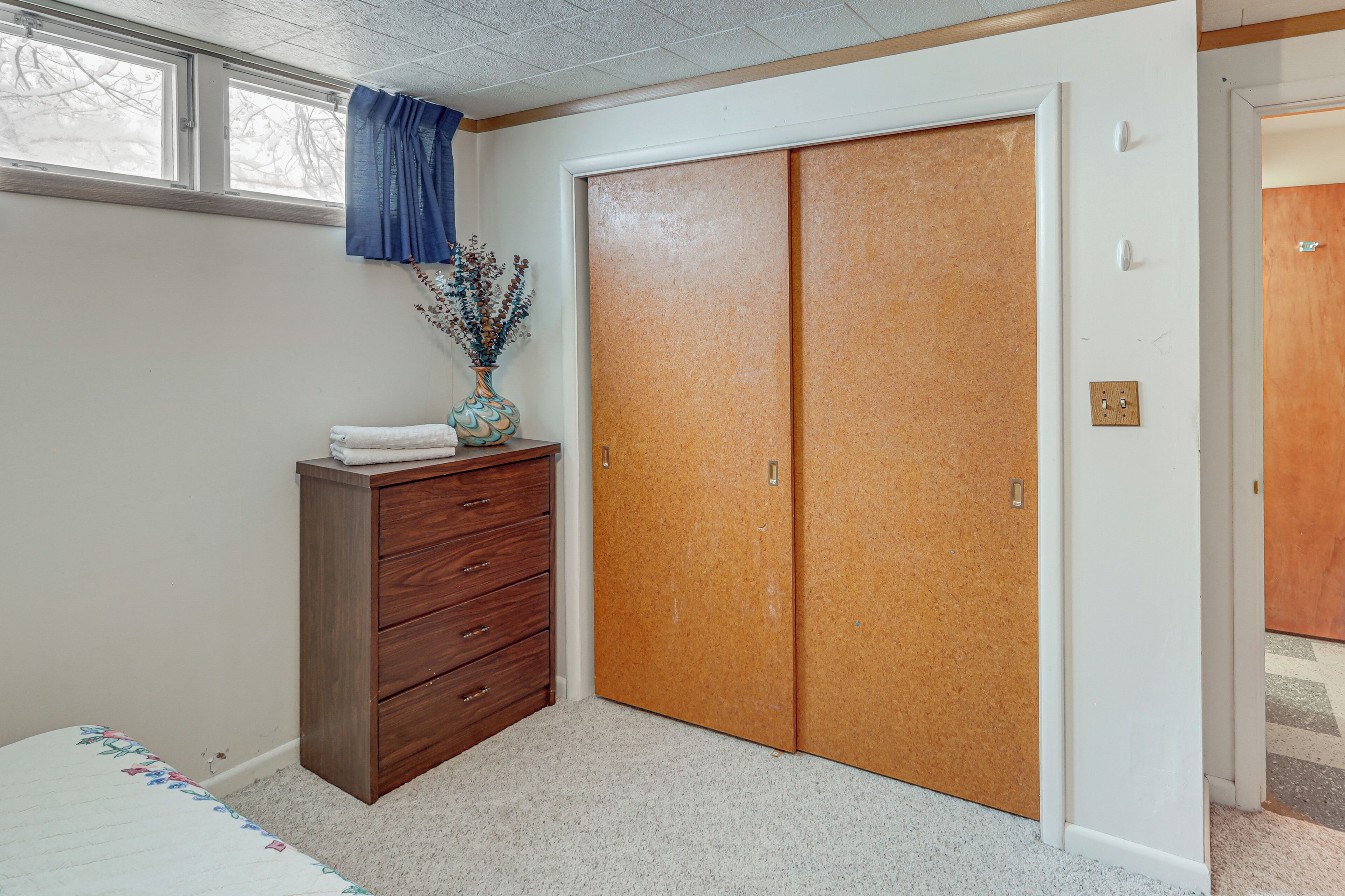 A bedroom with a wooden dresser, a closet with two doors, a window with blue curtains, and a vase with dried flowers on the dresser.